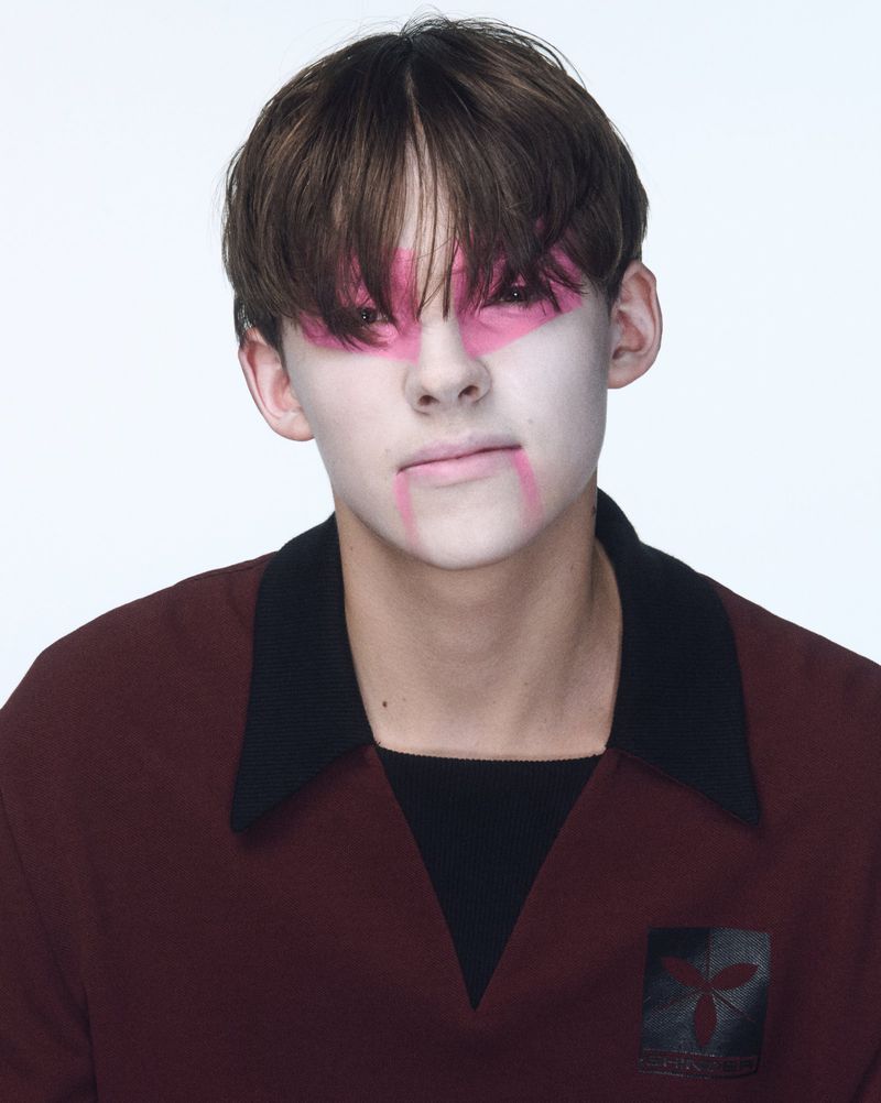 Portrait of a young adult with pink makeup around the eyes and mouth wearing a maroon shirt with a black collar against a white backdrop.