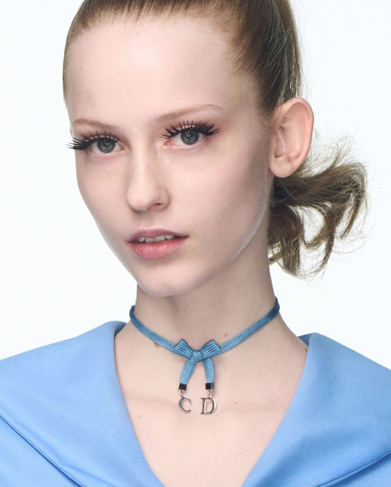 Close-up portrait of a fair-skinned woman with a blue ribbon choker necklace featuring the 'CD' logo, against a bright white backdrop.