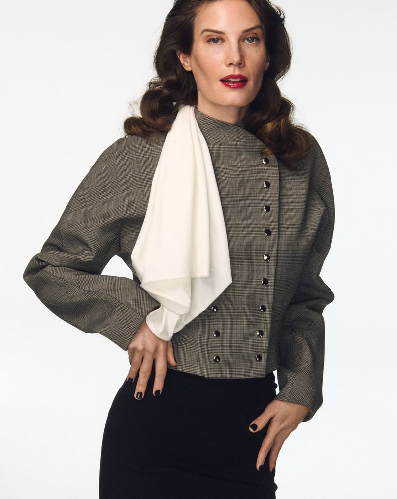 Studio shot of an adult woman in a grey double-breasted blazer and black skirt, posing against a white backdrop with hands on her hips.