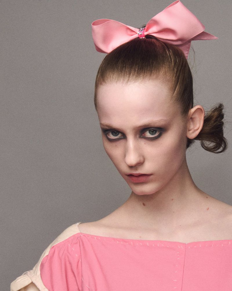 Woman portrait with pink bow hair accessory, wearing off-the-shoulder pink top in a studio setting with a neutral gray background.