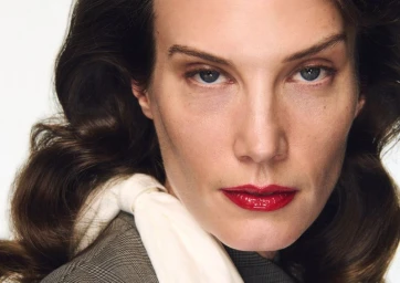 Close-up portrait of a woman with glossy red lips and dark wavy hair wearing a grey jacket against a white background.