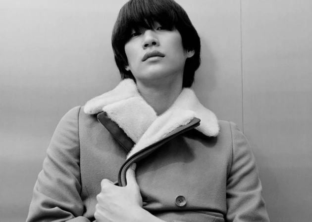 Black and white portrait of model Sanggun Lee wearing a shearling collar coat against a plain backdrop.
