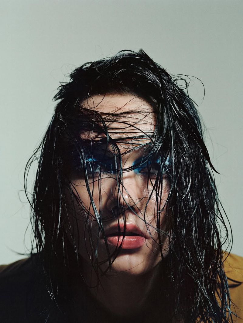 Close-up portrait of a young adult woman with dramatic blue makeup and wet black hair against a pale grey background. Beauty shot, editorial.