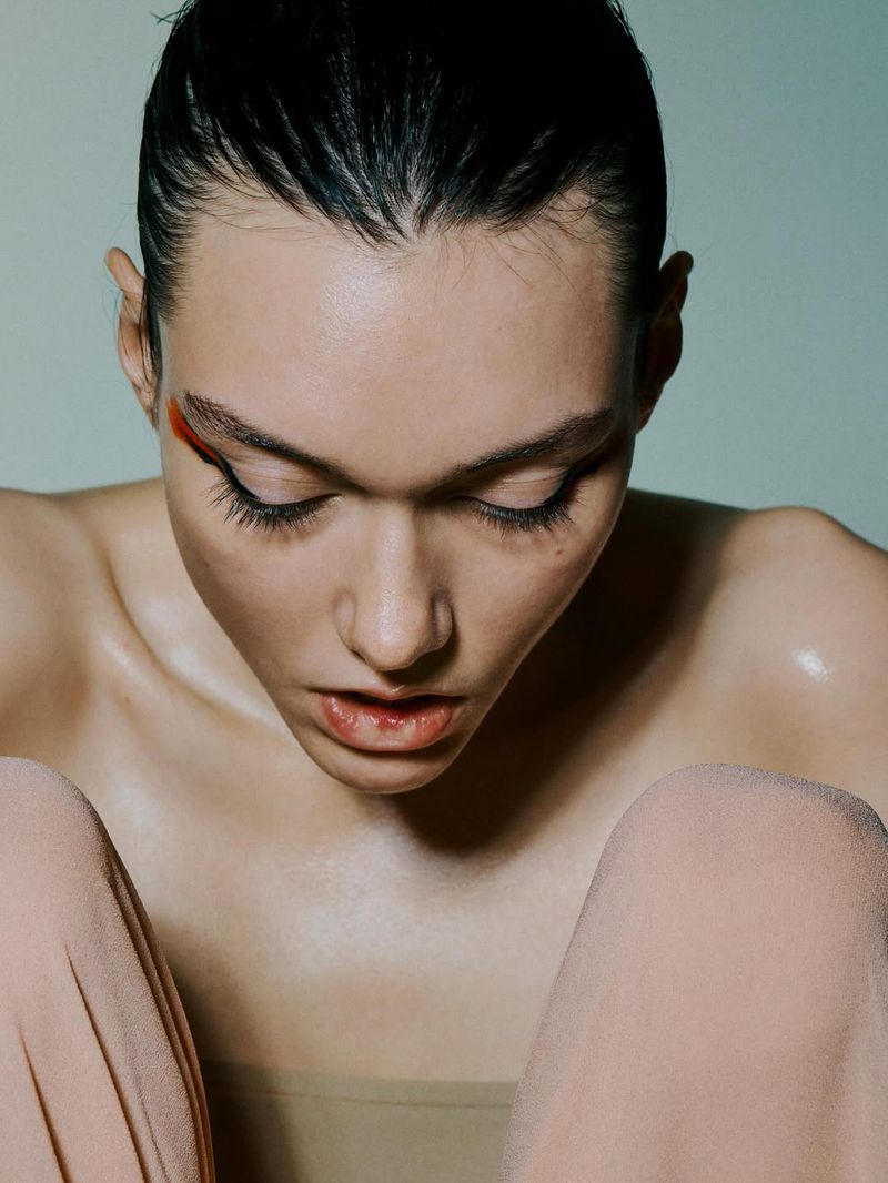 Close-up of a young woman with slicked-back hair and minimal makeup looking down, showing glowing skin and emphasizing natural beauty.