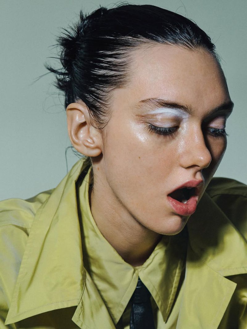 Portrait of a young adult woman with an open mouth, white eyeshadow, wearing a yellow jacket and black tie against a solid grey background.