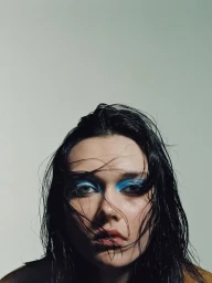 Close-up portrait of a woman with wet dark hair and vibrant blue eyeshadow against a plain background