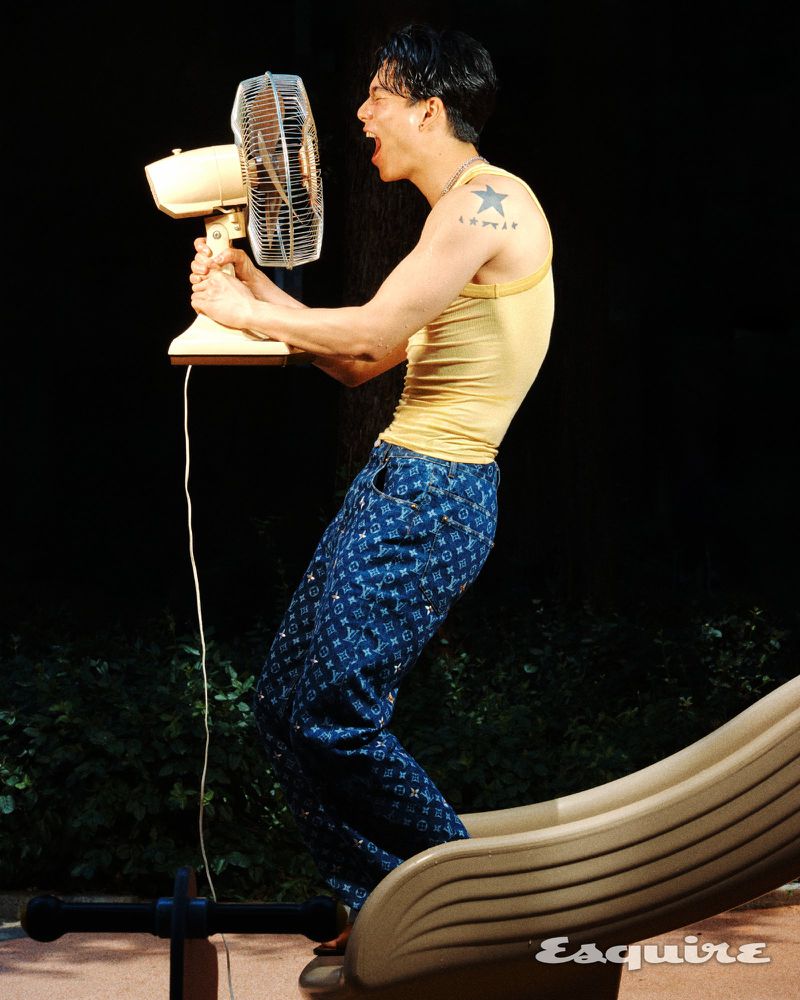 Model Joji Iwase holds an electric fan near his face while wearing a yellow tank top and patterned blue denim pants at night.