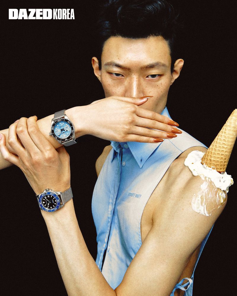 Close-up portrait of a male model wearing luxury watches and a light blue sleeveless shirt with an ice cream cone on his shoulder against a black background.