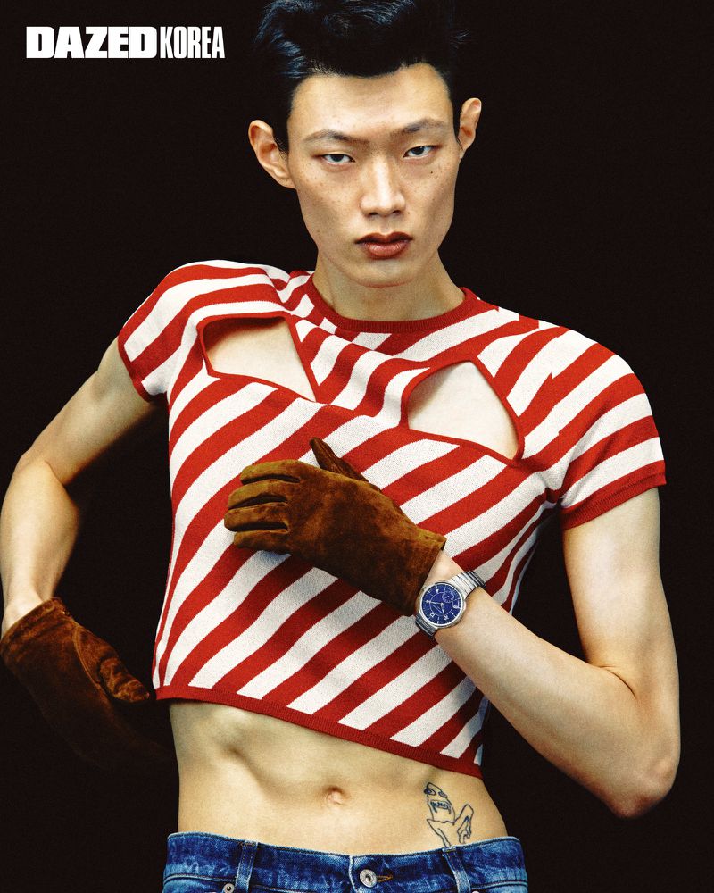 Model Hoyong Kim posing in a red and white striped cut-out crop top, brown suede gloves, and blue jeans against a black backdrop.