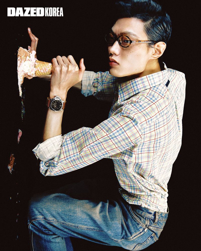 Man wearing eyeglasses, plaid shirt, and jeans holding an ice cream cone against a black background.