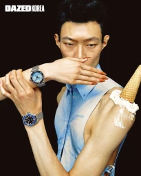 Close-up portrait of a male model wearing luxury watches and a light blue sleeveless shirt with an ice cream cone on his shoulder against a black background.