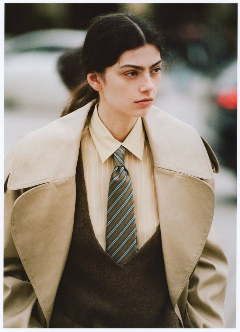 Portrait of a woman wearing a beige trench coat, brown sweater vest, striped button-down shirt, and striped tie in an outdoor setting.