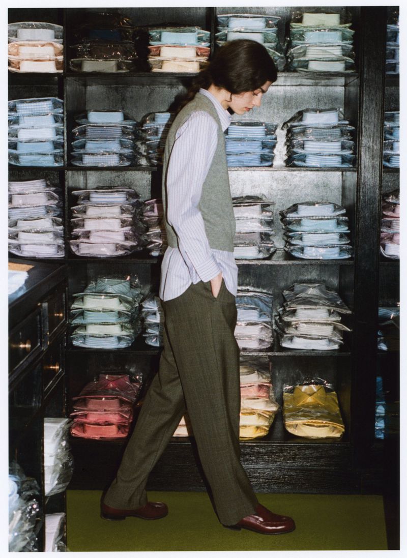 Adult male walks past shelves of folded shirts in a retail setting, wearing a button-down shirt, vest, trousers, and dress shoes.