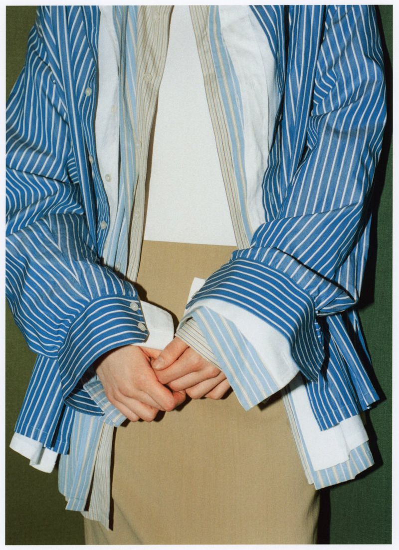 Unrecognizable person in beige skirt and blue-and-white striped button-down shirt with their hands clasped together, studio shot.