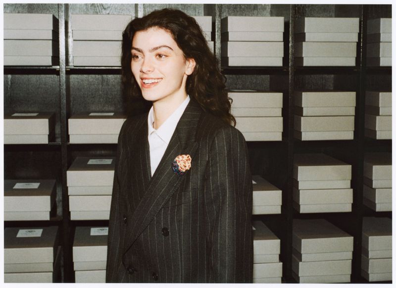Young woman smiles, wearing striped blazer with floral brooch, against a backdrop of stacked boxes on shelves in an indoor setting.