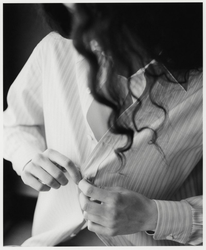 Black and white studio shot of an adult female buttoning a striped shirt, with long dark curly hair in a softly lit dark environment.