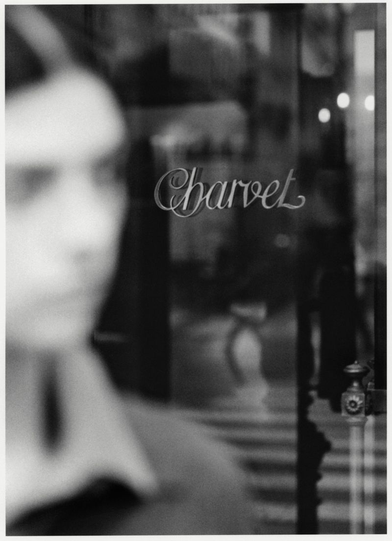Black and white image with blurry human face on the left, and Charvet logo on the glass of a building's storefront. 