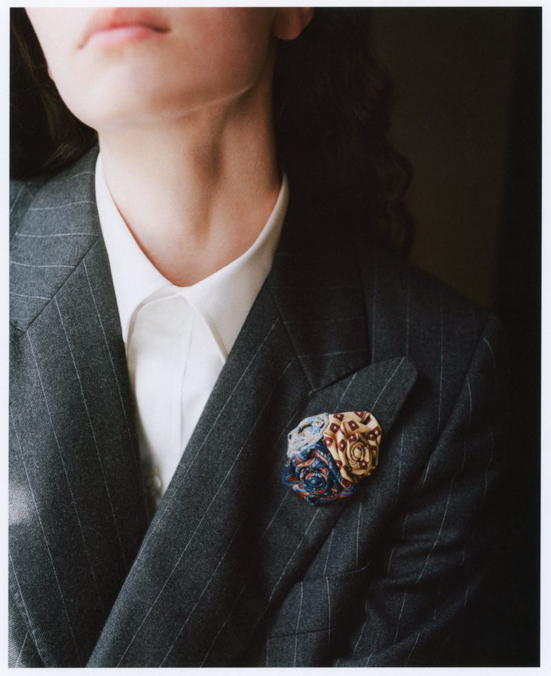 Woman in pinstripe suit jacket with floral pocket square, white collared shirt, dark hair, natural light, neutral background.