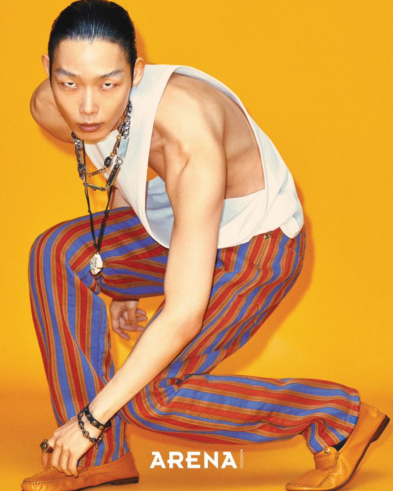 East Asian male model crouching dynamically in a white sleeveless top and striped red and blue trousers against a yellow backdrop.