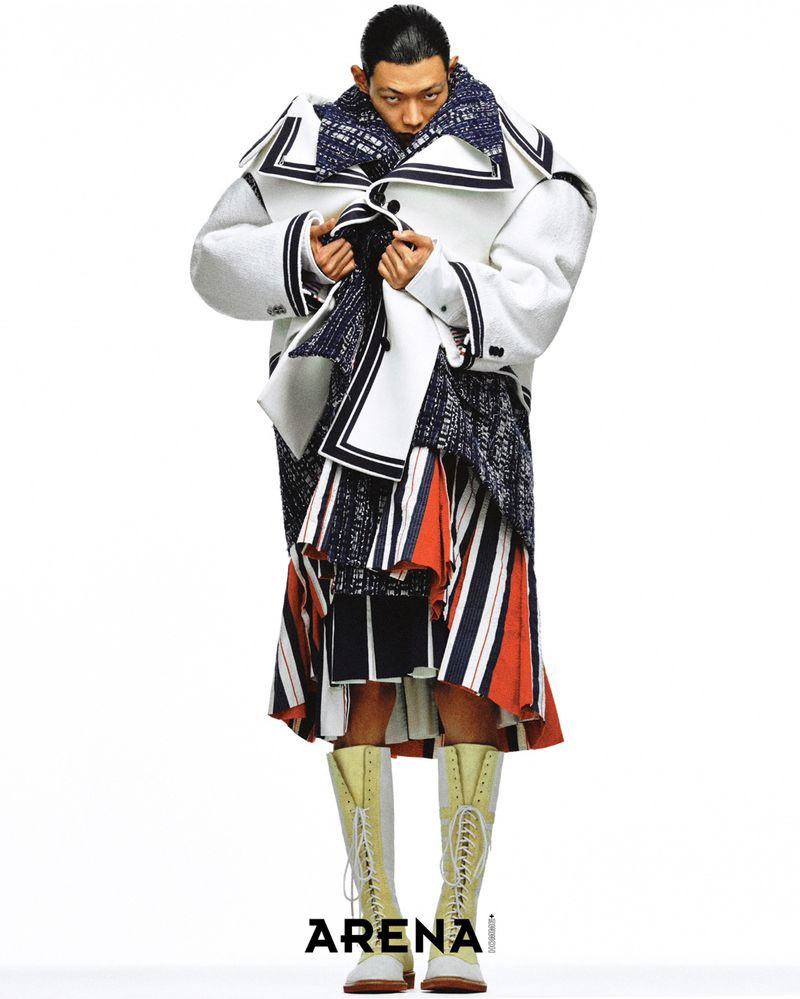 Male model posing in an oversized white sailor-collar coat, layered striped skirt, and knee-high lace-up boots against a white backdrop.