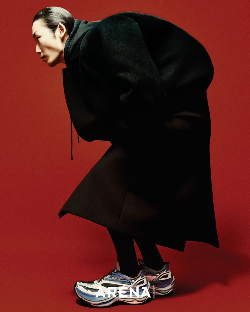 Model in an oversized black coat and futuristic sneakers bent forward against a bold red background.