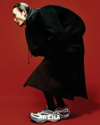 Model in an oversized black coat and futuristic sneakers bent forward against a bold red background.