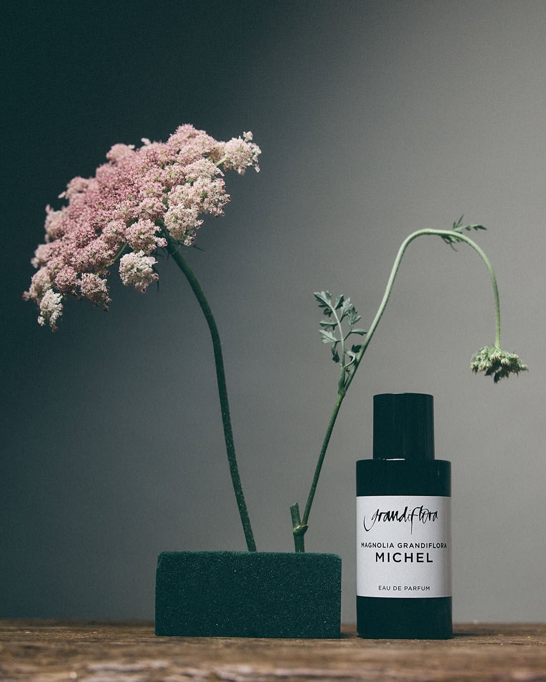 Still life featuring pink queen anne's lace in a foam block alongside a black Grandiflora Michel perfume bottle on a wood surface.