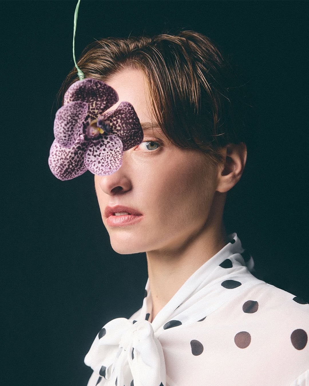 Close-up studio portrait of a young adult woman with an orchid covering one eye, wearing a sheer polka dot blouse against a dark backdrop.