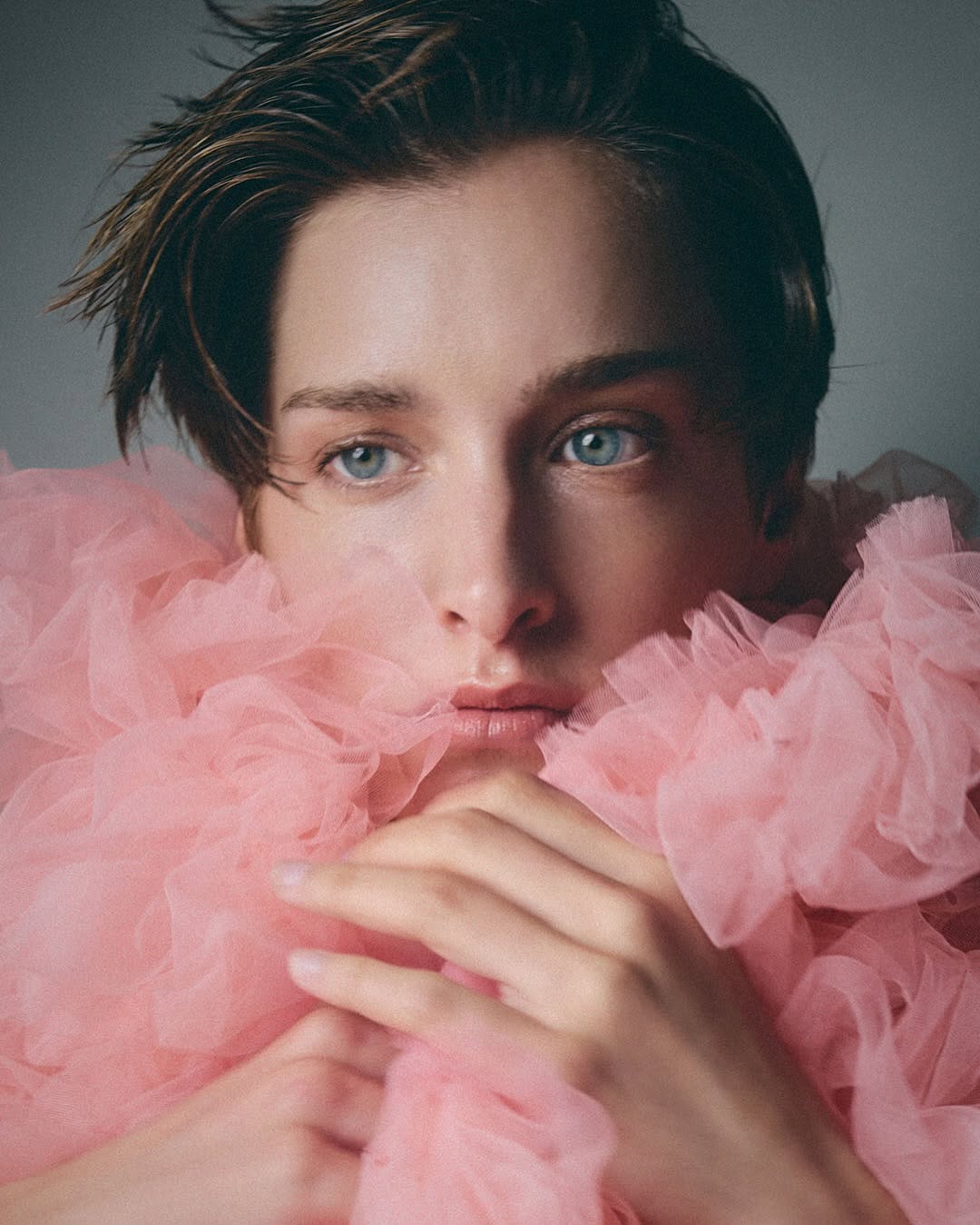 Close-up portrait of an adult with light skin, blue eyes, and brown hair, wearing a pink tulle garment, against a gray background.