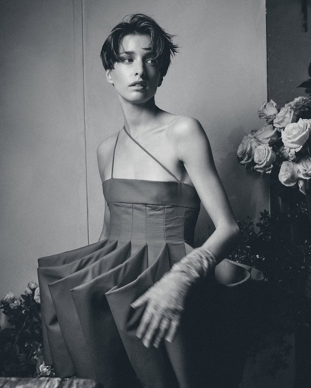 Monochrome fashion shot of a woman with short hair wearing a sculptural red dress, standing in a studio with floral accents.