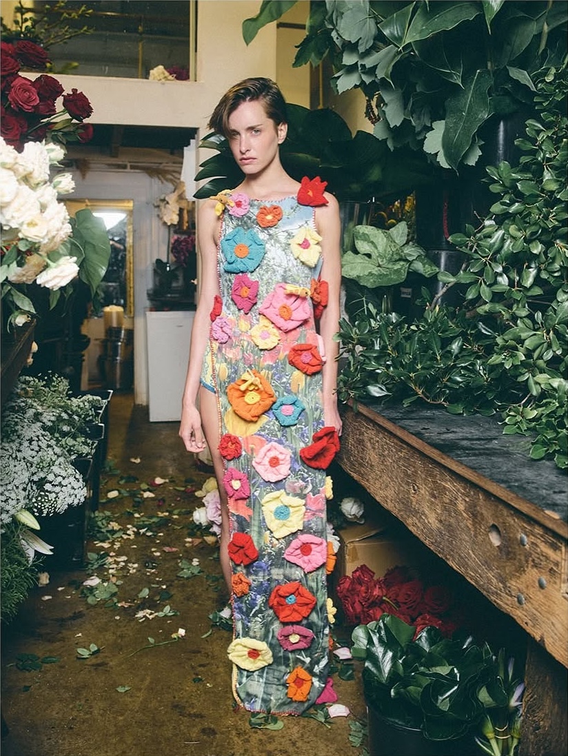 A woman stands in a floral-themed gown amid lush greenery and cut flowers, creating a striking blend of fashion and natural beauty.
