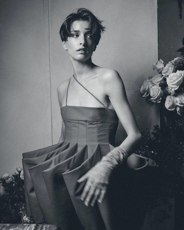 Model with short dark hair in a structured strapless dress beside a large arrangement of roses in a monochromatic editorial portrait