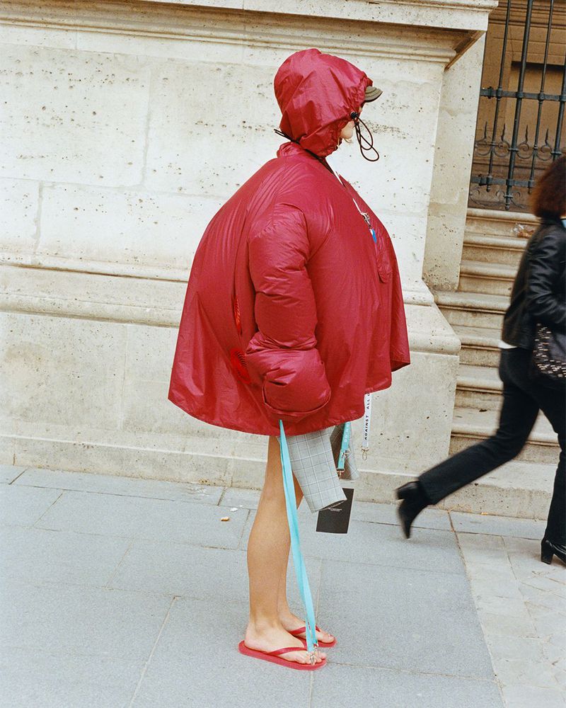 Person in oversized red puffer coat and flip flops walking on sidewalk next to a building with pedestrian in background.