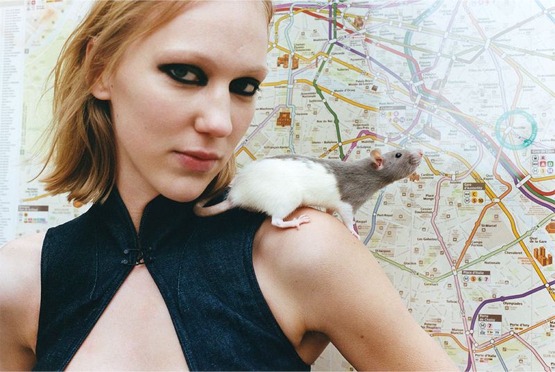 Young woman with heavy eye makeup poses with a fancy rat perched on her shoulder against a Paris metro map backdrop.