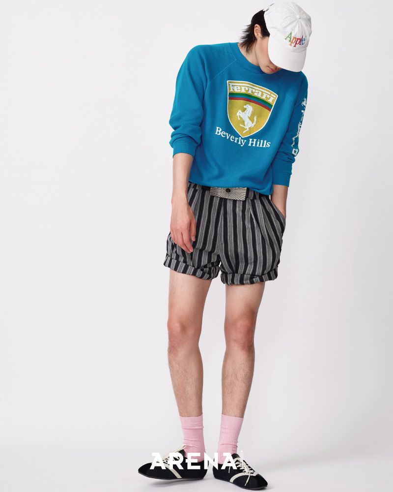 Model Jinyeong Pak in a blue graphic sweatshirt, striped shorts, pink socks, and white cap against a plain white studio backdrop.