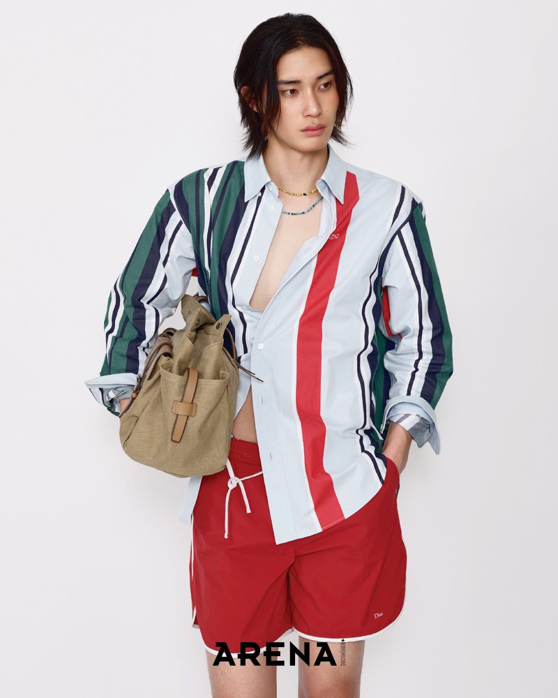 Model Jinyeong Pak wearing an unbuttoned striped shirt and red shorts with a tan canvas bag against a white backdrop.