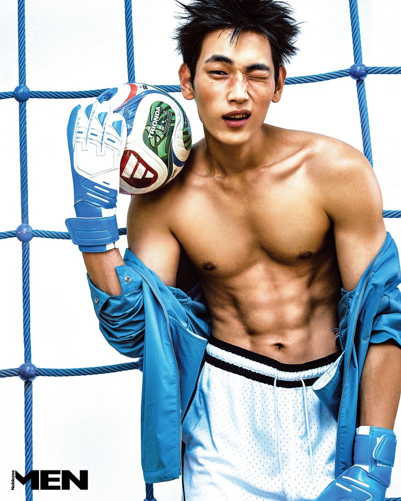 Athletic East Asian man winking while holding a soccer ball, wearing blue goalkeeper gloves and white shorts against blue netting.