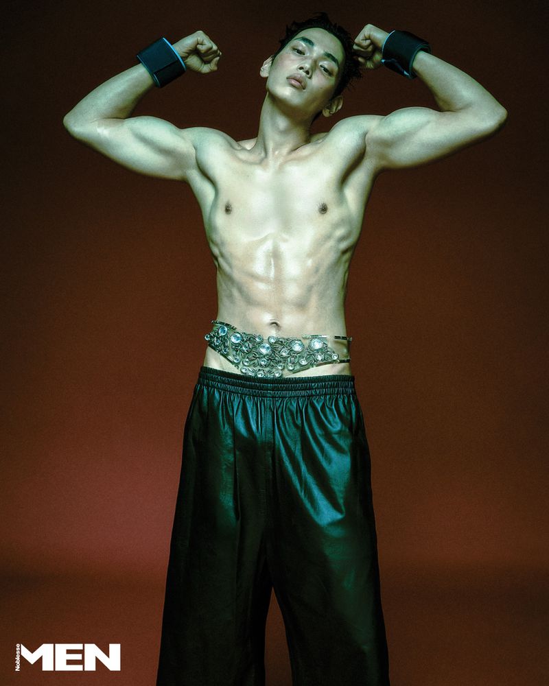 Shirtless man flexing his arms wearing black wide-leg leather trousers and a crystal-embellished belt against a dark red backdrop.