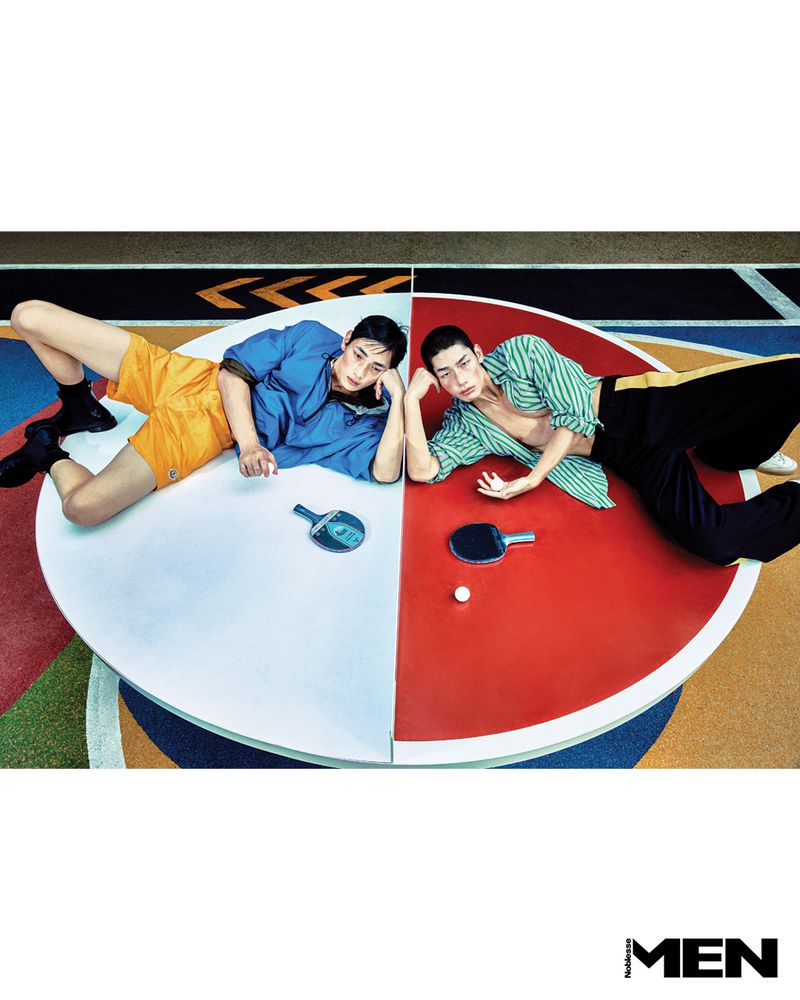 Two male models lying on a circular red and white table tennis table with ping pong paddles and balls on a colorful court surface.