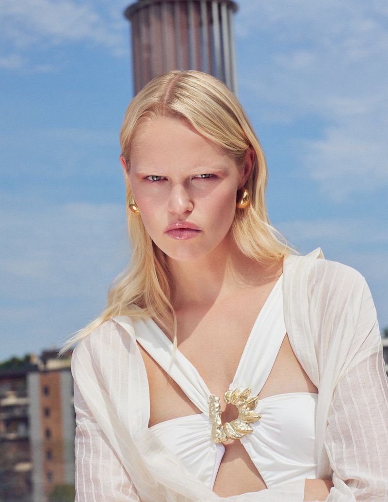 Model Roos Schreiner posing in a white cutout top with a gold brooch and earrings under a blue sky for Grazia Italy.