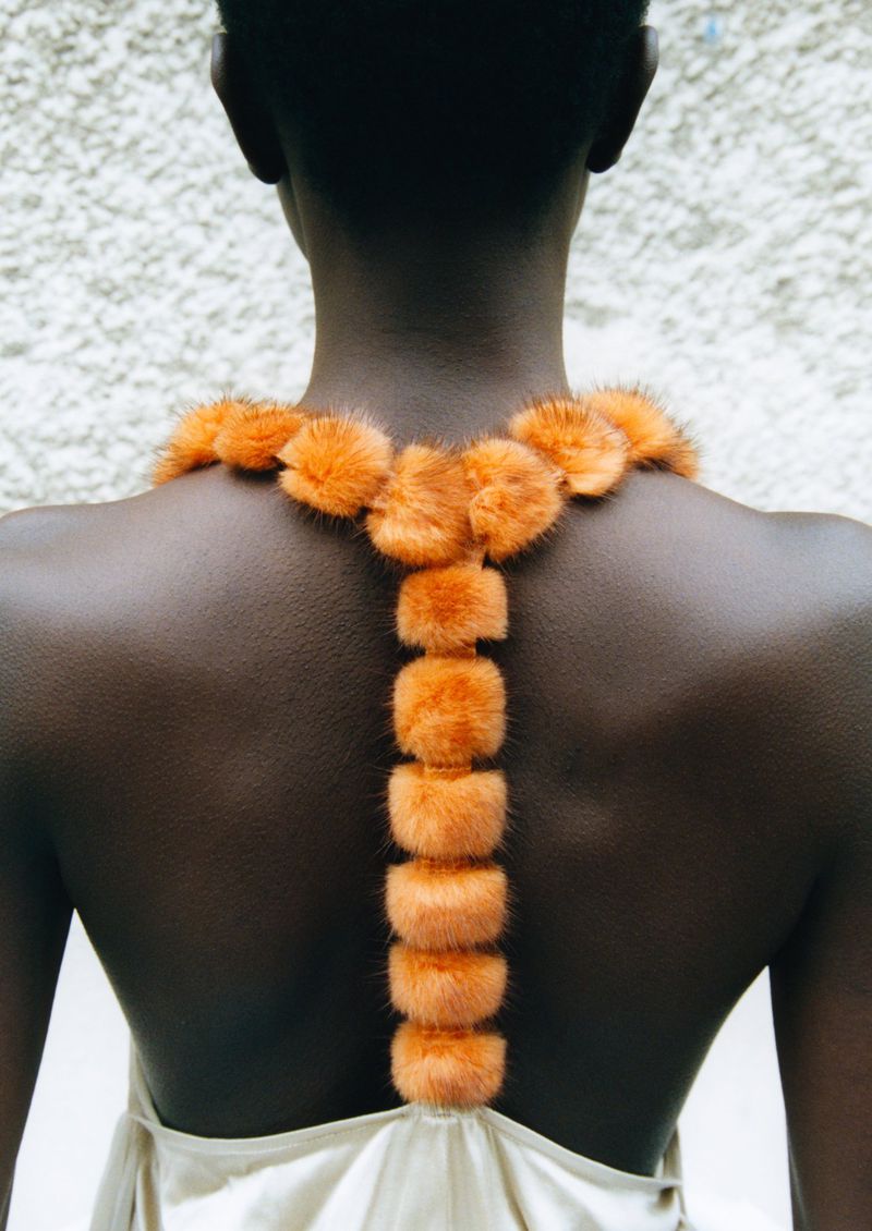 Back view of a Black female adult wearing a light-colored dress and orange fur-like neckpiece, standing against a textured wall.