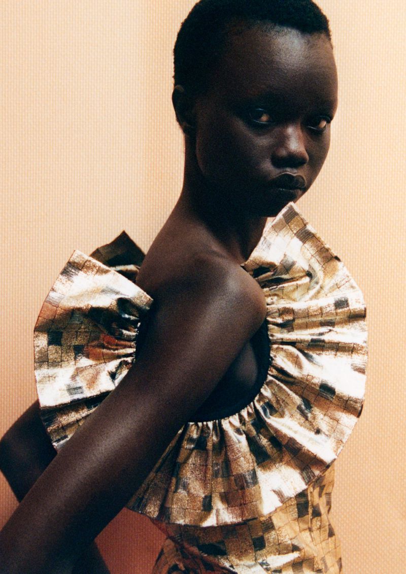 Studio portrait of a Black female fashion model in a gold dress with a large ruffled collar against a warm, peach-toned background.