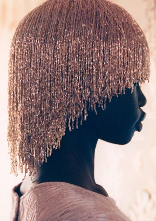 Profile shot of a woman wearing an intricately beaded headpiece, draped fabric top, and minimal makeup against a blurred background.