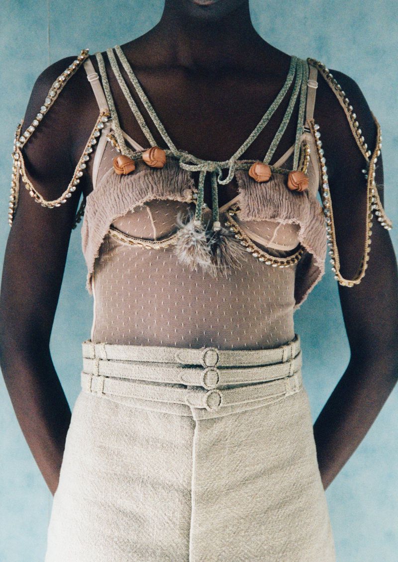 A slender, dark-skinned model poses in a cropped linen pant and embellished bra top, complemented by a soft blue background in a fashion editorial.