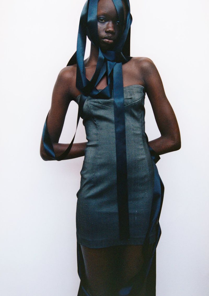 Dark-skinned woman in a denim dress poses in a studio with dark blue ribbon accents, presenting a fashion editorial shot against a white background.