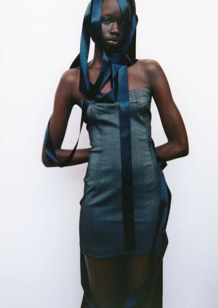 Dark-skinned woman in a denim dress poses in a studio with dark blue ribbon accents, presenting a fashion editorial shot against a white background.