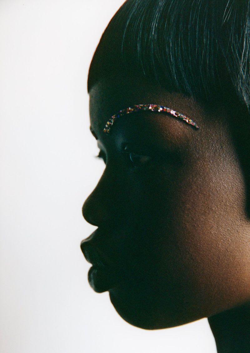 Close-up profile portrait of a woman with dark skin, a jewel embellishment on her eyebrow, and short dark hair against a white backdrop.