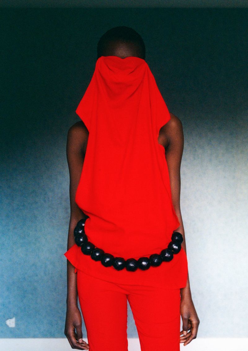 A slim Black woman in a vibrant red outfit poses against a textured teal wall, her face obscured by red fabric, complemented by a black beaded belt.