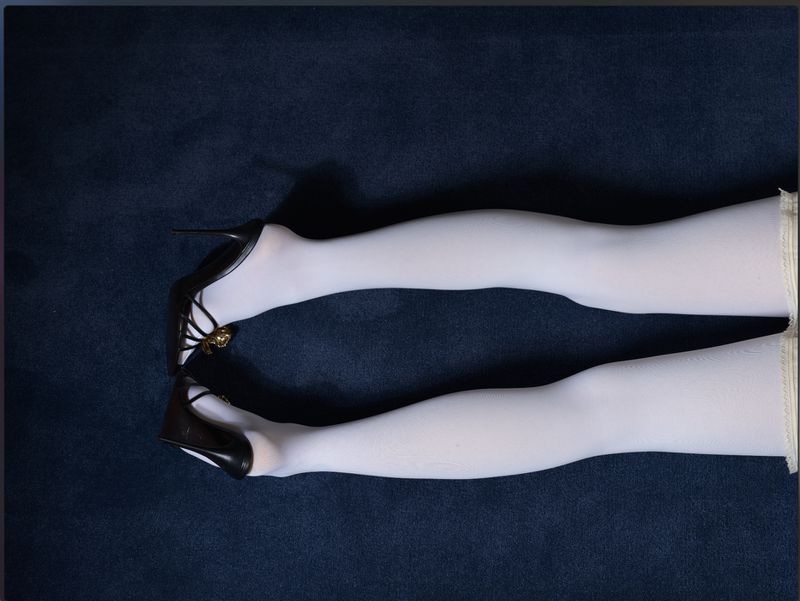 Close-up fashion shot of a woman's legs in white tights and black high heels on a dark blue textured fabric background in a studio setting.