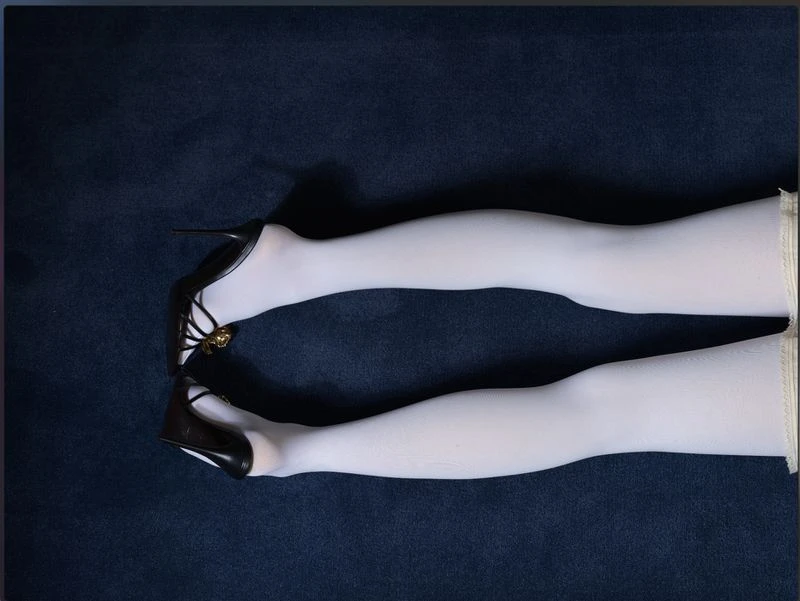 Close-up fashion shot of a woman's legs in white tights and black high heels on a dark blue textured fabric background in a studio setting.